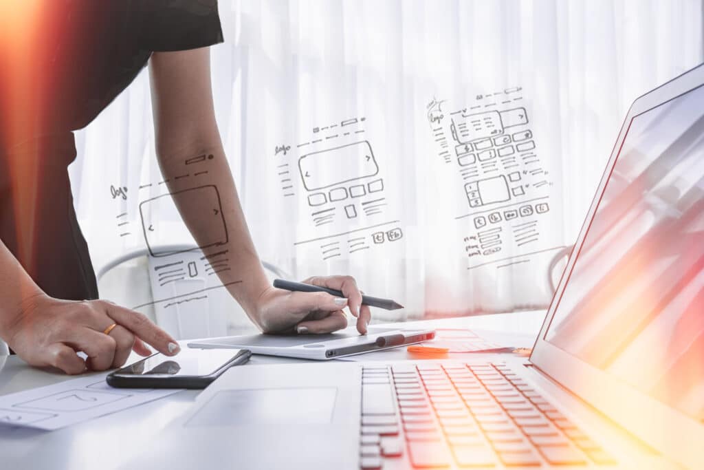 A man leans over a desk with laptop, phone and drawing tablet holding an e-pencil. Superimposed are illustrations and plans for website UX and SEO.