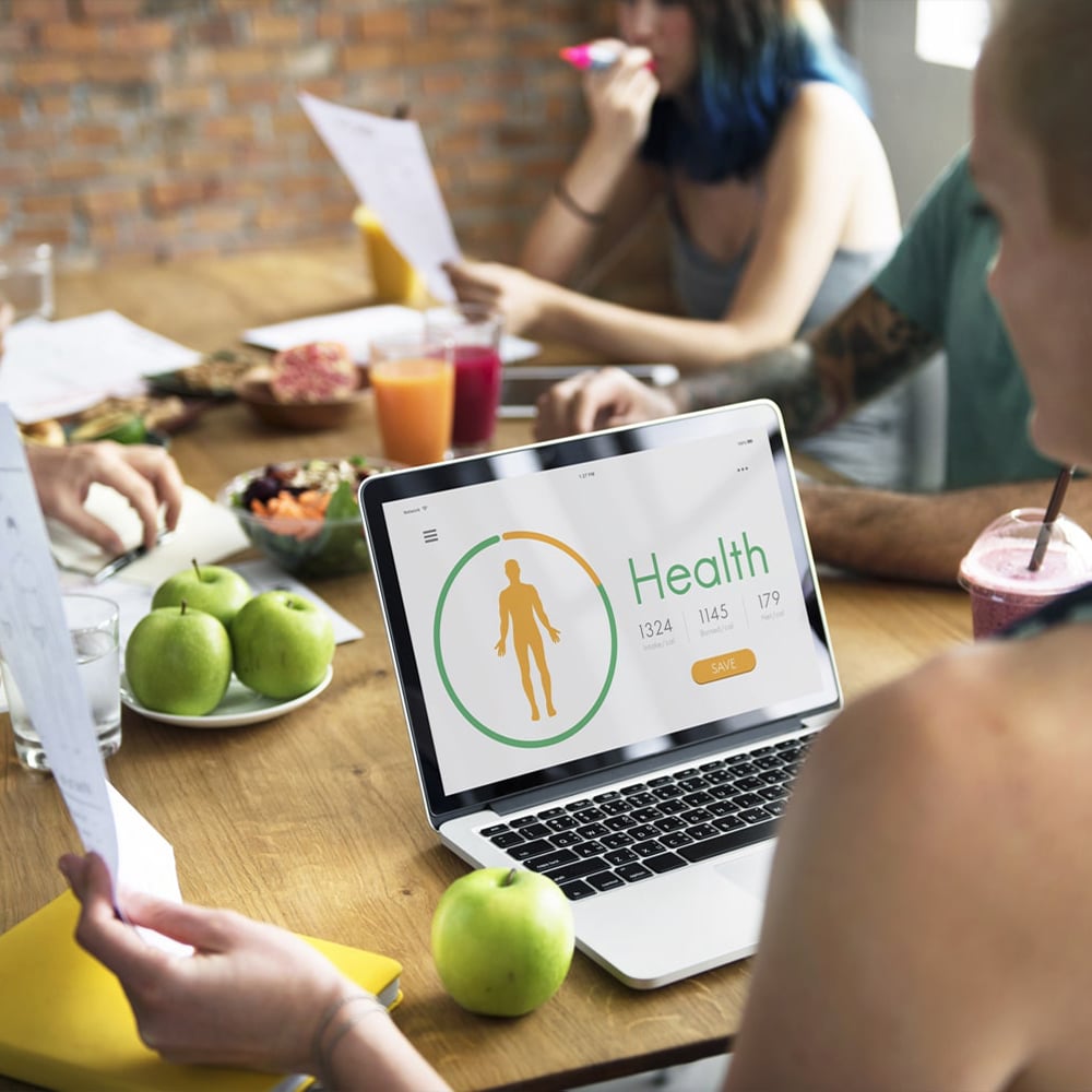 A woman attends a meeting, on the desk in front of her is an areray of healthy food and fruit as well as a laptop showing a health report. Square pic