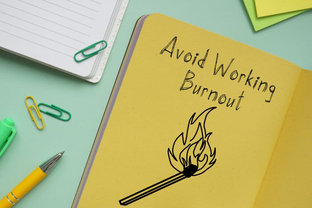 A notebook on a messy desk has a drawing of a burning matching and the words 'Avoid Working Burnout' written on it