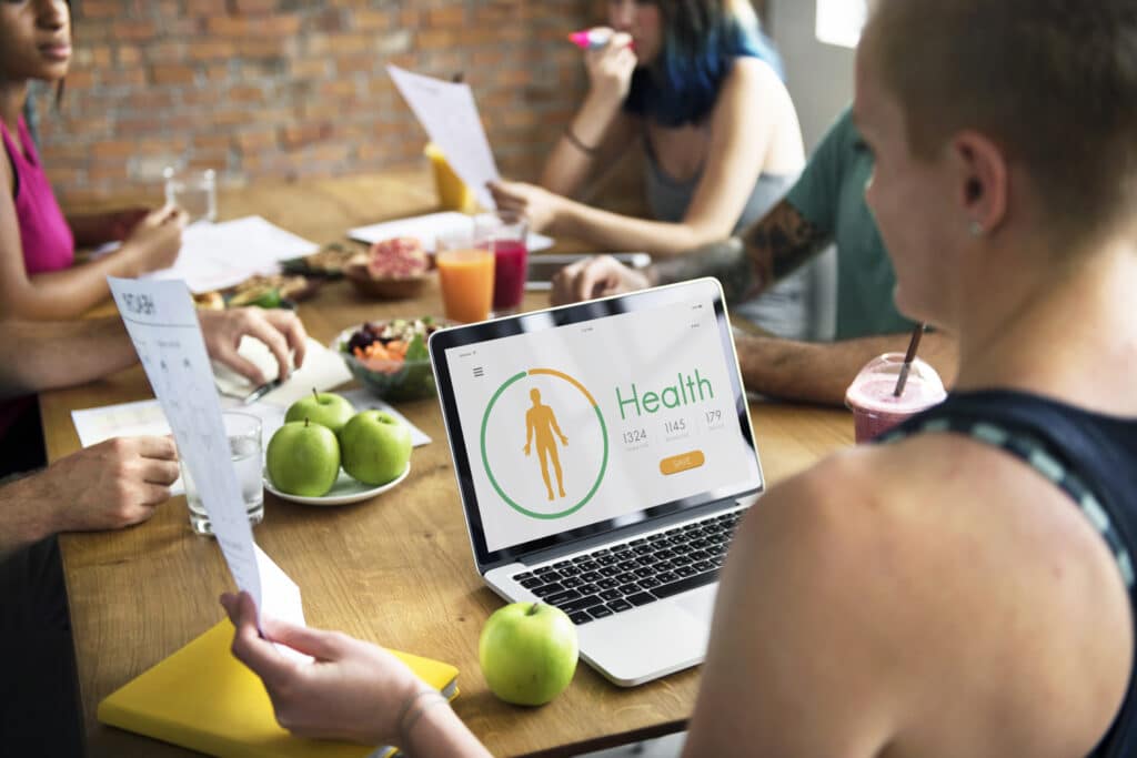 A woman attends a meeting, on the desk in front of her is an areray of healthy food and fruit as well as a laptop showing a health report.