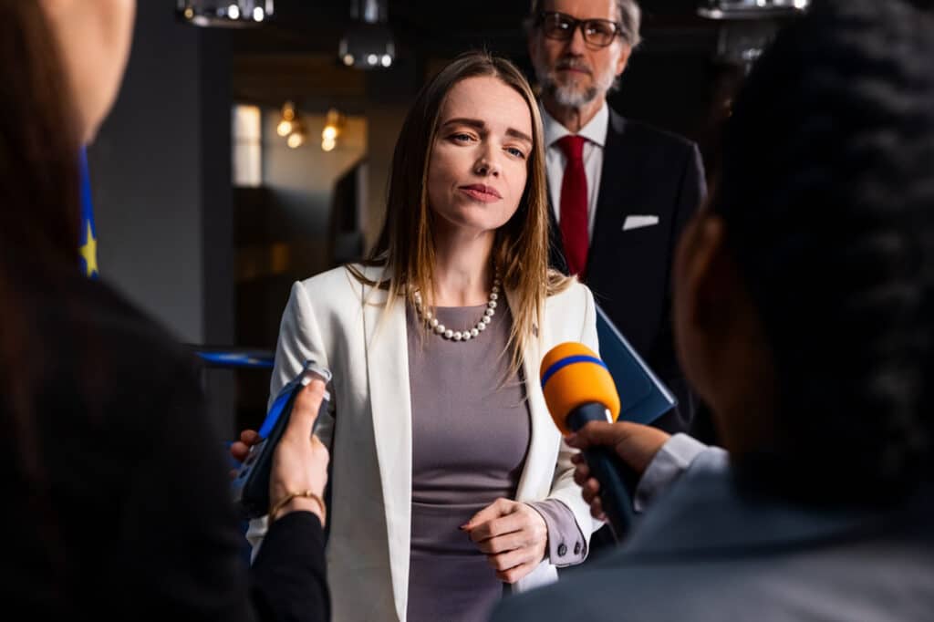 A woman answers questions for the press as journalists hold microphones in front of her.