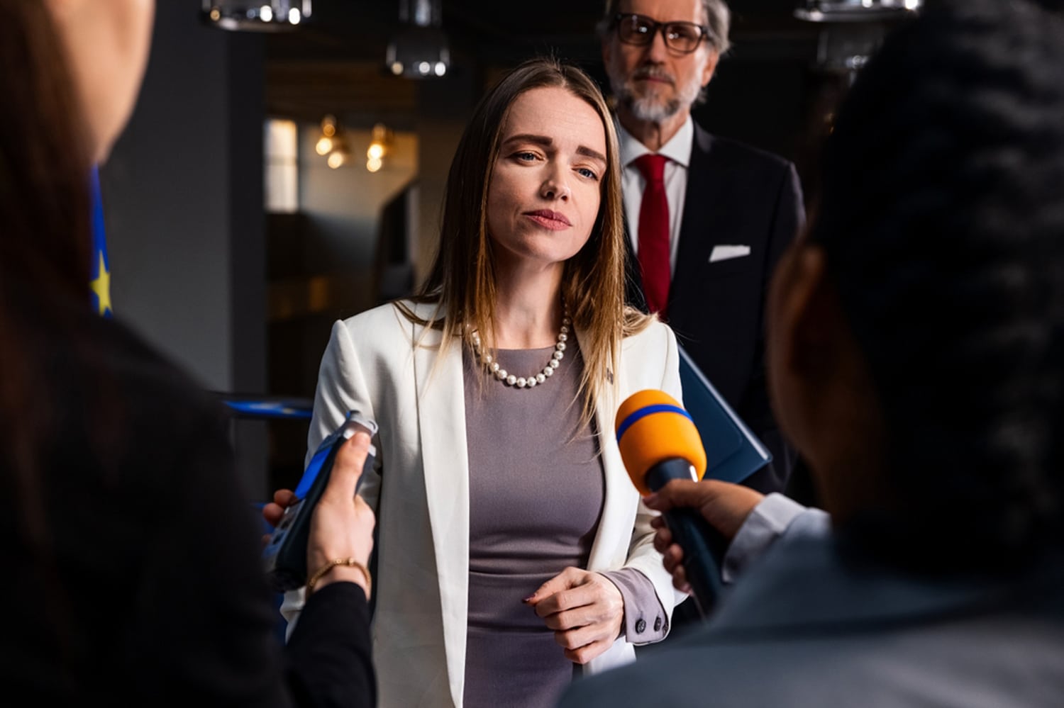 A woman answers questions for the press as journalists hold microphones in front of her.